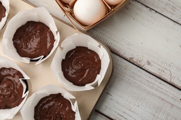 Chocolate dough in cupcake tray and ingredients for muffins on white wooden table, flat lay. Space for text
