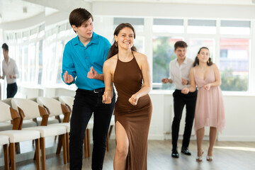 Fototapeta premium Smiling graceful girl in fitted brown dress and stylish guy in blue shirt and black trousers rehearsing jazz routine in dance studio, performing traveling steps and snapping fingers