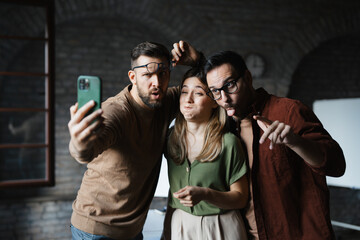 Group of three diverse colleagues making funny faces and taking a selfie in a modern office.