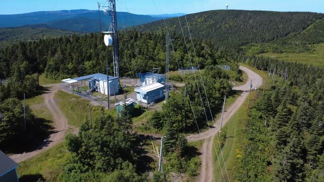 A verdant mountain range is surrounded by communication antenna towers, a Doppler radar, and a wind farm on the horizon. Matapedia Valley, Quebec, Canada, 2025.