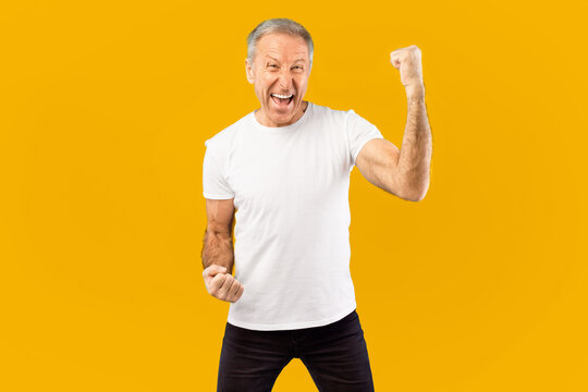 A man shows excitement by raising his fist and smiling. He wears a plain white shirt and black pants. The background is bright yellow, creating a strong and energetic atmosphere.