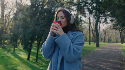 Woman enjoys warm drink in park during morning while wearing coat and ear muffs surrounded by trees...