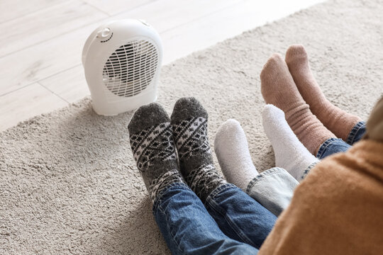 Family with lack of heating warming feet near electric fan heater at home
