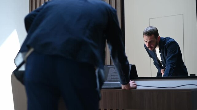 Caucasian programmer working on his laptop at a modern office, standing in front of the desk, coding, checking his phone, and looking at his reflection in the mirror with concentration