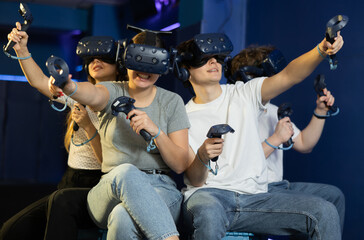 in computer club, group of four friends wearing virtual reality glasses and with joysticks are sitting and playing dynamic game with modern graphics.