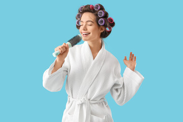 Young woman in bathrobe with hair curlers and brush singing on blue background