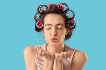 Young woman with hair curlers blowing kiss on blue background, closeup