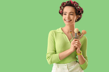 Young woman with hair curlers and makeup brushes on green background