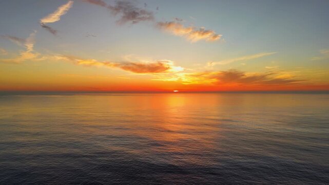 Aerial drone flying backward over the Atlantic ocean during a vivid red, orange, and yellow sunset in Portugal, showing colorful sky reflections and calm water from an elevated perspective