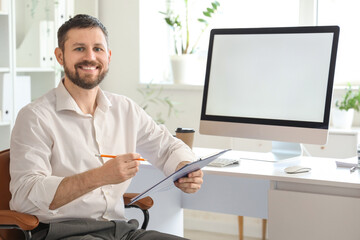 Naklejka premium Young businessman working with clipboard and blank computer at table in office