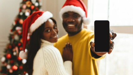 Fototapeta premium A couple wears Santa hats and stands in front of a decorated Christmas tree. They smile and hold a smartphone, sharing a joyful moment during a holiday celebration.