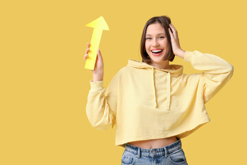 Happy young woman with paper arrow pointing at something on yellow background