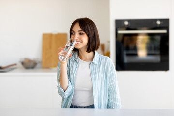 A young woman with dark hair smiles as she drinks a glass of water in a bright, modern kitchen. Natural light fills the space, creating a cheerful atmosphere.