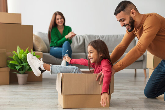 Excited Arab family celebrates moving day in their new apartment. The father plays with his daughter, sliding her in a cardboard box while the mother smiles from the couch, creating joyful memories.