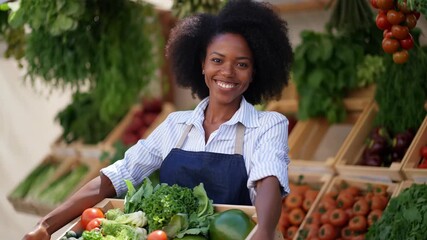 Young woman smiling at camera while holding a wooden crate overflowing with fresh organic vegetables and fruit, standing at a bustling farmers market stall, waist-up portrait