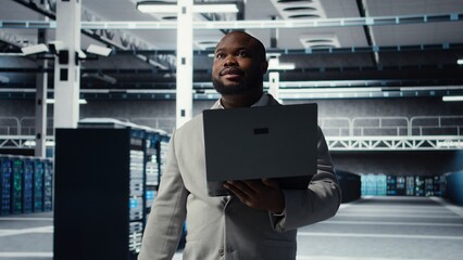 Technician walking in data center, configuring storage devices for minimal latency in AI workloads. African american employee using device in server farm to build neural networks, camera A