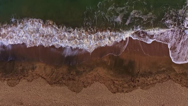 Warm sunlight glimmers on the ocean waves as they roll onto a tranquil sandy beach. The tide creates foam that gently hugs the shore, offering a peaceful atmosphere at dusk.