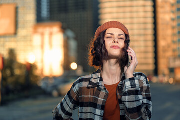 Fototapeta premium Woman on phone in urban street at golden hour, beanie and plaid shirt, confident smile and candid lifestyle portrait conveying authenticity, mindful living and emotional storytelling.