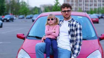 Young man and little girl in eyeglasses posing near red car outdoors. Guy showing thumb up. Family, transport, trip, safety and parenthood concept. Slow motion