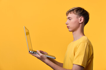 Youth in a yellow shirt holding an open laptop against a bright yellow background, focused on the screen, illustrating online learning, digital exploration and tech focus.