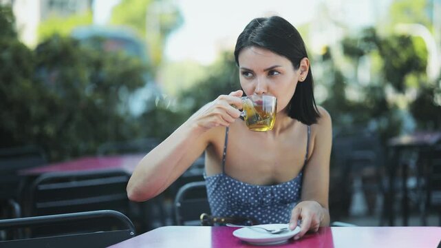 Elegant young woman in summer dress enjoying tea while sitting alone in open-air cafe. High quality 4k footage
