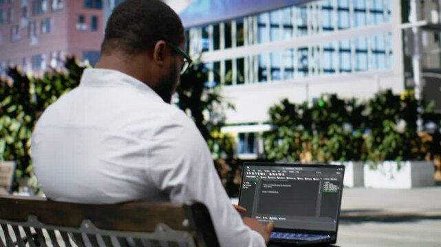 African american guy works on programming code on terminal window, sitting on a bench downtown and working on IT with his laptop in sunlight. Professional attitude in a dynamic city life. Camera B.