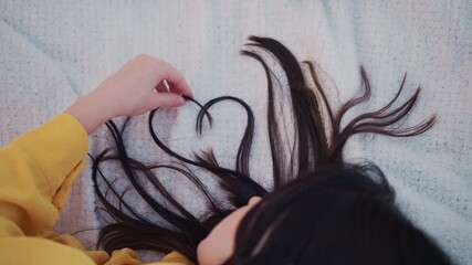 asian woman lying on knit blanket, long dark hair arranged into heart shape as fingers gently guide strands, yellow sweater sleeve visible, soft pastel tones, cozy lofi vibe, intimate quiet moment,
