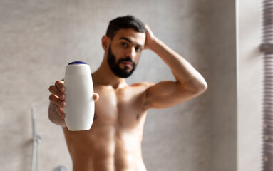 Fototapeta premium A confident Arab guy stands in a stylish bathroom, holding a shampoo bottle. He poses while raising one arm and showing off his toned physique, promoting a male cosmetic product.