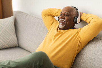 Fototapeta premium An elderly man, wearing headphones, relaxes on a comfortable sofa in his living room. He smiles as he enjoys listening to music, creating a peaceful atmosphere.