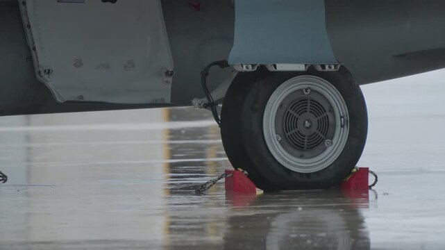 Military aircraft landing gear wheel secured with chocks on wet runway reflecting rain and ground operation safety measures