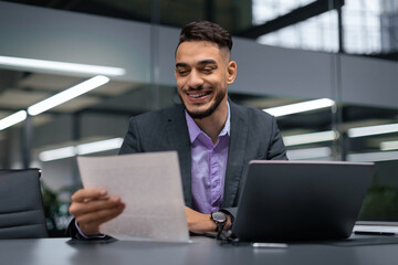 A man sits at a desk in a modern office, smiling as he reads a document in his hands. A laptop is open in front of him, and bright light fills the room from large windows.