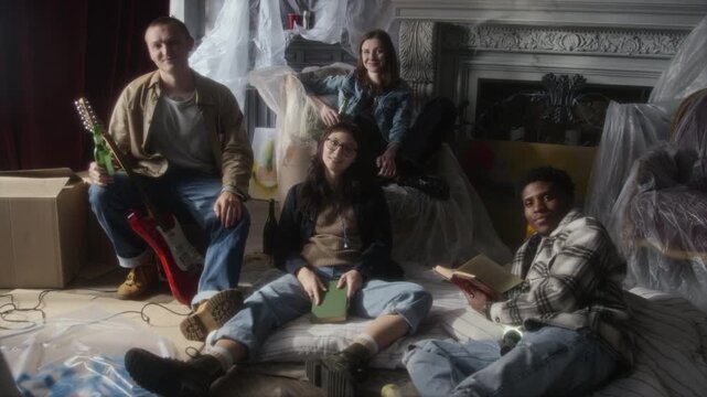 Handheld full length shot of eclectic group of young people sitting on floor in abandoned house all looking at camera while hanging out together, soft focus