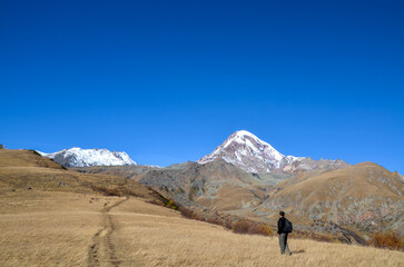 Hiker with a backpack stands on a grassy, golden-brown hillside trail, looking toward the snow-capped summit of Mount Kazbek under a clear blue sky in the Greater Caucasus Mountains