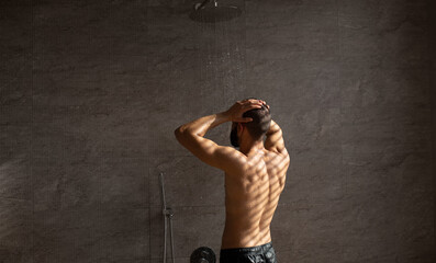 Muscular guy stands under warm water in a stylish bathroom, following his daily hygiene routine. He enjoys the refreshing shower while maintaining his body care.
