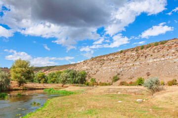 Arid landscape with arid, tiered rock formations rising sharply from valley floor containing stream and patches of green riparian vegetation. Calm river water and rugged sedimentary layers of hillside