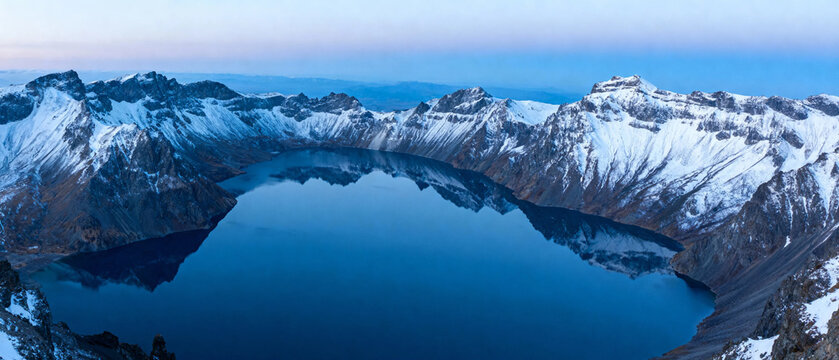 Snow-capped mountain range surrounding a calm, deep blue crater lake at dawn - Powered by Adobe