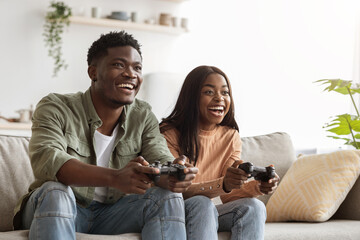 Obraz premium A joyful black couple is sitting on their couch, holding game controllers and enjoying a fun gaming session. Their expressions show excitement and happiness as they bond in their living room.