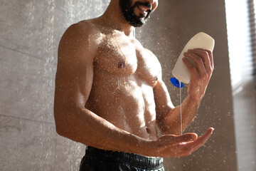 Muscular young man stands in a contemporary bathroom, pouring shampoo onto his palm. Water falls...