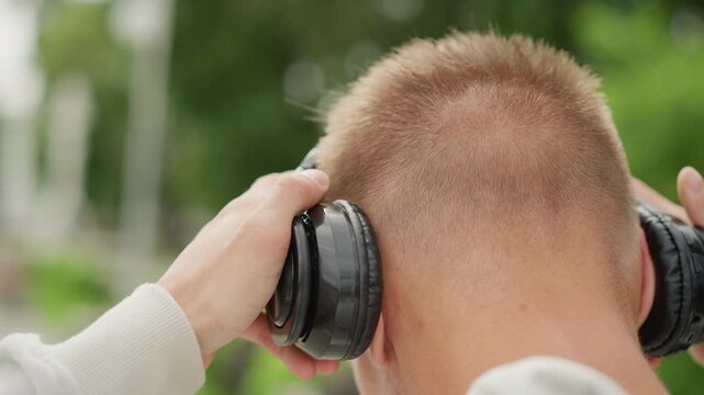 park path side profile testing headphones while walking. Casual daytime scene with greenerylined promenade, listener rotating earcup and gesturing with hand, subtle sunlight through clouds frames