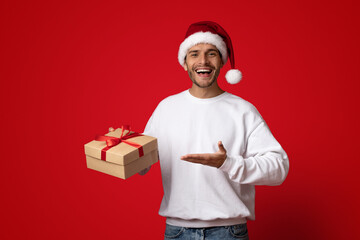 A joyful man wearing a Santa hat stands against a vibrant red background, holding a beautifully wrapped gift box with a ribbon while smiling cheerfully.