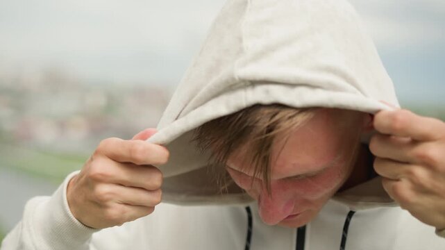 young white male soaked in rain wearing drenched hoodie, hair plastered to forehead, hood tugged down, eyes closed with emotion, city skyline blurred, rain droplets on fabric, vulnerable cathartic
