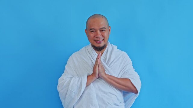 Happy Muslim man in ihram greeting with hand together isolated on blue background