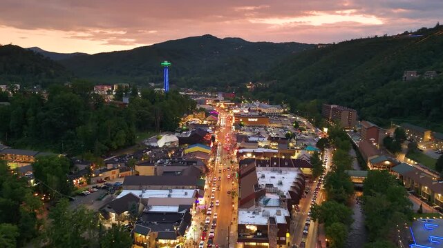 Aerial landscape of evening at Gatlinburg city center with busy tourist streets, main Parkway filled with shops and restaurants, and scenic Smoky Mountains in the background.