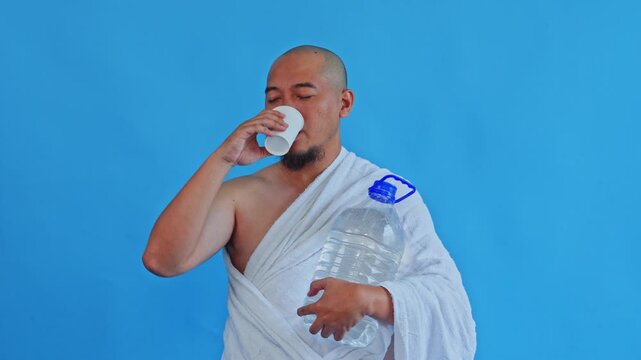 Bald Asian pilgrim in Ihram drinking Zamzam water isolated on blue background