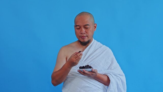 Muslim pilgrim wearing Ihram eats dates and gives thumbs-up isolated on blue background