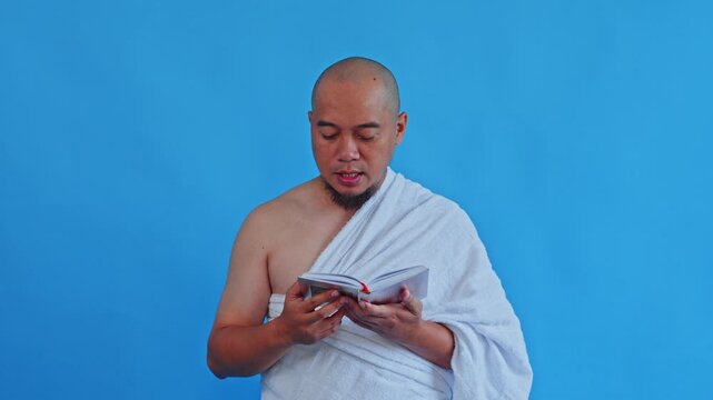 Muslim pilgrim in Ihram reading small prayer book on blue background