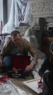Vertical shot of young man setting record on record player while sitting on floor in abandoned house in secret hideout with group of people