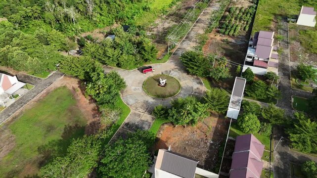 The view from above an urban roundabout featuring a central statue, surrounding green space, power lines, and neatly lined houses. 