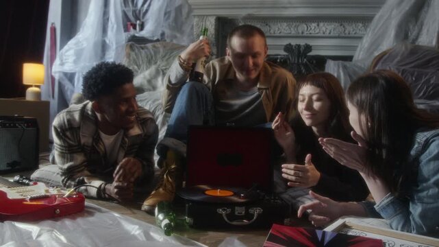 Full length front view shot of diverse group of young people listening to music with retro record player while sitting on floor and drinking beer hanging out together in abandoned house