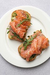 Tasty bruschettas with salmon, arugula and avocado on light grey table, top view